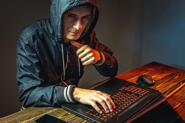 A hacker in a hood with a phone is typing on a laptop keyboard in a dark room The concept of cybercrime fraud and identity theft