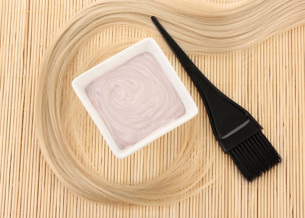 Hair dye in bowl and brush for hair coloring on beige bamboo mat closeup