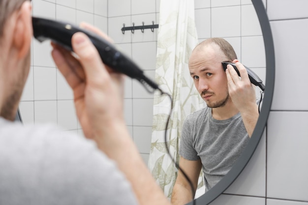 Hair loss problem A young man standing against mirror in bathroom cutting hair using hair clipper