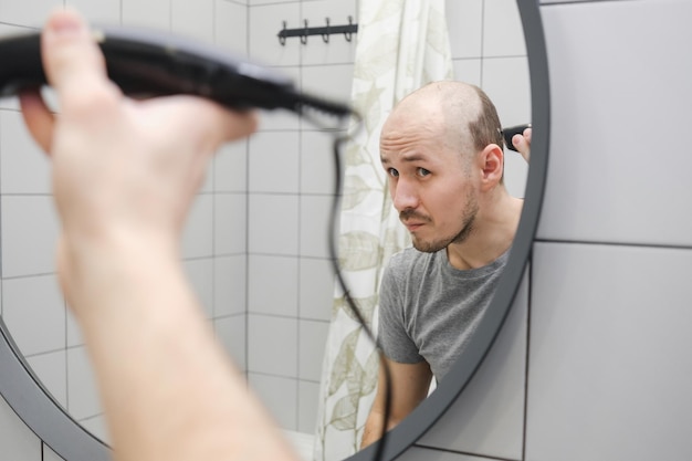 Hair loss problem A young man standing against mirror in bathroom cutting hair using hair clipper