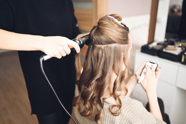 Hair stylist prepares beautiful young woman for event, makes curls hairstyle with a curling iron for client in beauty salon