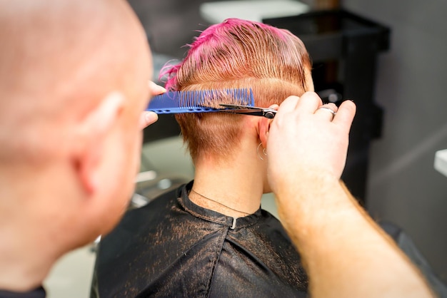 Haircut of dyed short pink wet hair of young caucasian woman by a male hairdresser in a barbershop