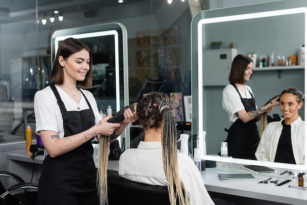 hairdresser and clientbeauty salontattooed hair stylist doing hair of cheerful woman with braids