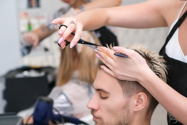 Hairdresser cuts the hair of a young man with blond dyed hair with scissors in a beauty salon. Professional hair care.