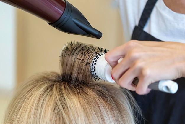 Hairdresser dries wet hair, makes hair styling a hairdryer and comb to a blonde client