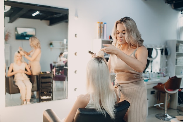 Hairdresser female making hair extensions to young woman with blonde hair in beauty salon. Professional hair extension.