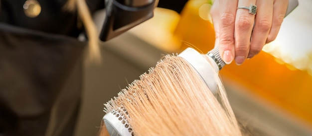 Photo hairdresser hand drying blond hair with a hairdryer and round brush in a beauty salon.