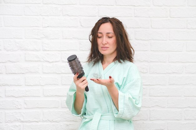 Hairloss concept worried young woman holding bunch of fallen hair in hand while standing near white