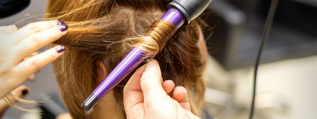 The hairstylist makes curls hairstyle of long brown hair with the curling iron in hairdresser salon close up