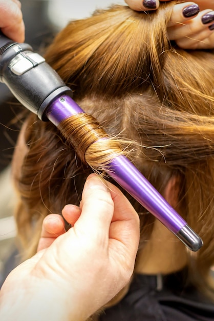 The hairstylist makes curls hairstyle of long brown hair with the curling iron in hairdresser salon close up