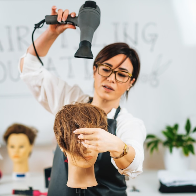 Photo hairstylist with the hair dryer in hands explaining hairstyling techniques using mannequin head