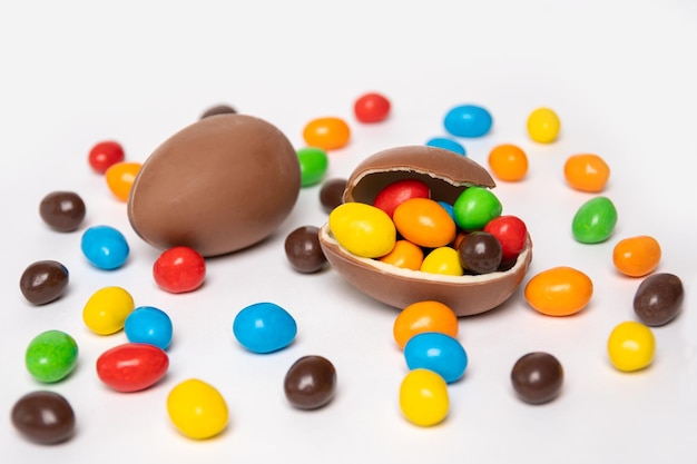 Half of a cracked chocolate egg filled with multicolored candies covered with an egg shell on a white background with multicolored candies Treats for children