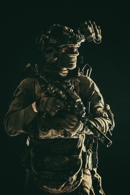 Half length low key portrait of elite commando fighter professional mercenary hiding identity behind mask glasses standing in darkness with mini submachine gun in hands wearing nighvision device