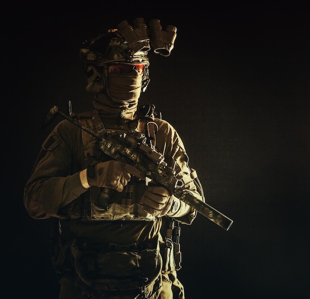 Half length low key portrait of elite commando fighter professional mercenary hiding identity behind mask glasses standing in darkness with mini submachine gun in hands wearing nighvision device