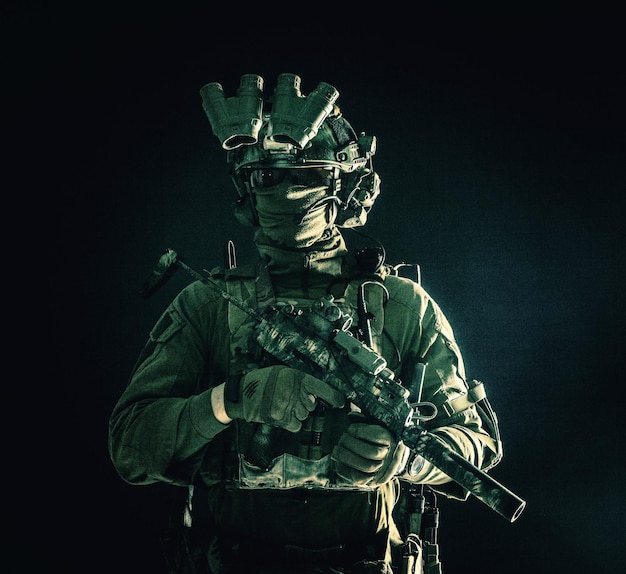 Half length low key portrait of elite commando fighter professional mercenary hiding identity behind mask glasses standing in darkness with mini submachine gun in hands wearing nighvision device