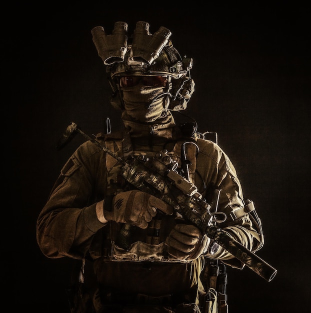 Half length low key portrait of elite commando fighter professional mercenary hiding identity behind mask glasses standing in darkness with mini submachine gun in hands wearing nighvision device