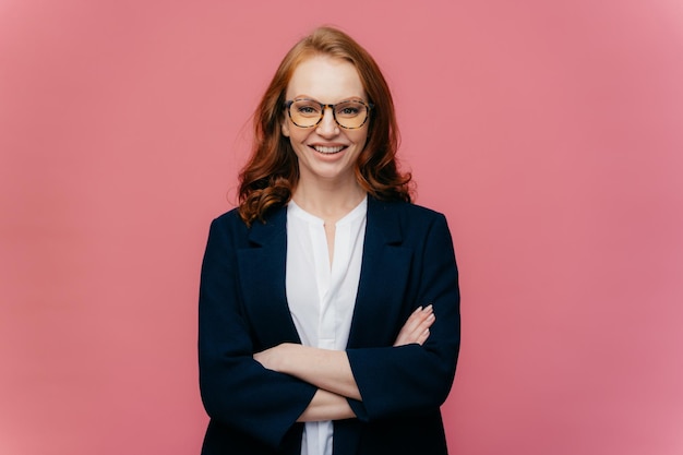 Half length shot of ginger business lady satisfied after conference, keeps arms folded, wears optical glasses, elegant black suit, models over pink background. Satisfied female manager poses indoor