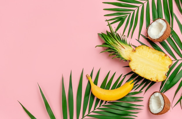 Half of pineapple, coconuts and banana with palm leaves on pink background. Copy space