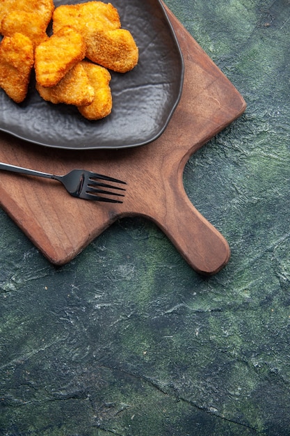 Half shot of chicken nuggets on a black plate and fork on wooden cutting board on the right side on dark surface with free space