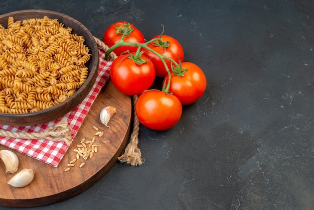 Half shot of raw pastas in a brown bowl on red stripped towel garlics rice on round wooden board tomatoes rope