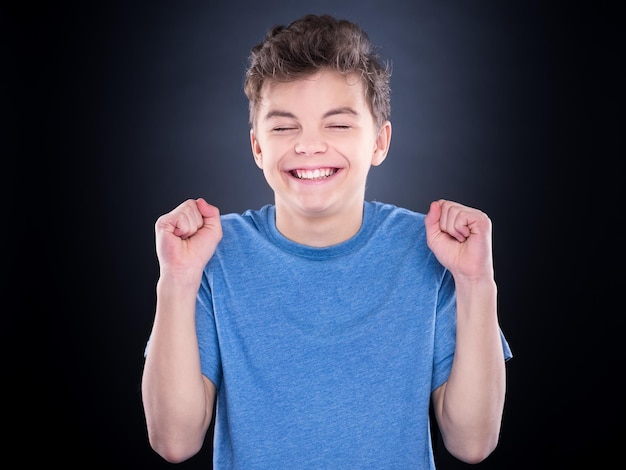 Halflength emotional portrait of caucasian teen boy Victory screaming teenager wearing blue tshirt on black background Funny winner child shouting with his hands up