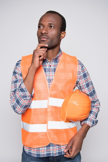 Halflength shot of thoughtful construction worker in a vest and a hardhat touching his chin and