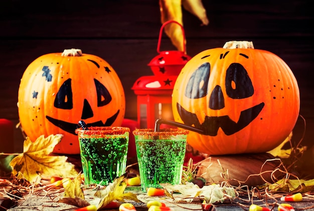 Halloween composition with festive green drink and smiling drinking pumpkins with sweet corn straw and fallen leaves on dark background selective focus