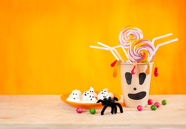 halloween concept. A glass with sweets and straws and a plate with cakes on an orange background