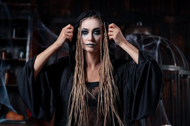 Halloween concept. Witch portrait close up with dreadlocks looking camera dressed black hood standing dark room with cage on background Caucasian female necromancer