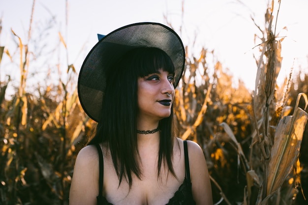 Halloween costume witch girl portrait in a cornfield at sunset. Beautiful serious young woman in witches hat with long black hair and dark lips.