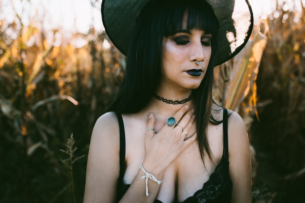 Halloween costume witch girl portrait with close eyes in a cornfield at sunset. Beautiful sad and serious young woman in witches hat with long black hair and dark lips.