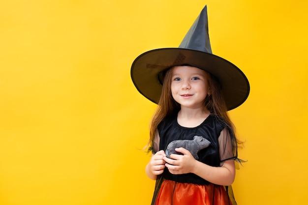 Halloween. A funny girl in a witch costume holds a rat. Yellow background, space for text. Emotional portrait. High quality photo