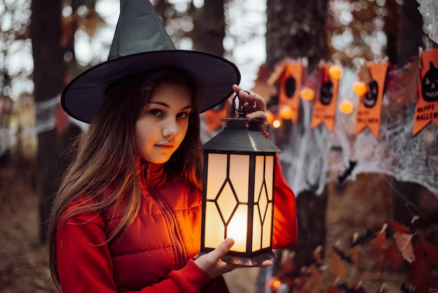 Halloween girl in witch hat with lantern in her hands in the forest