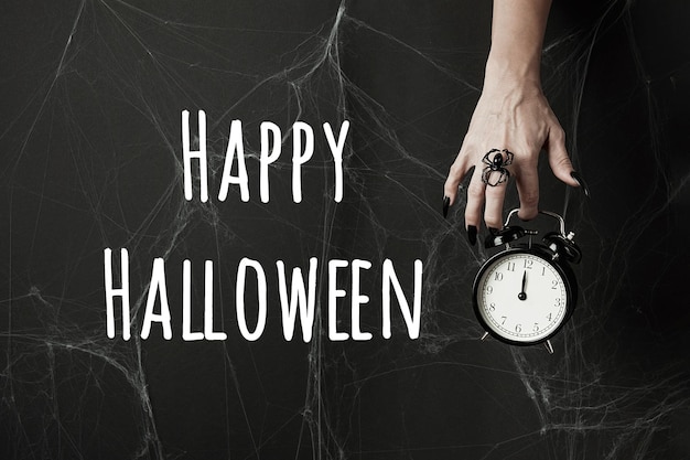 Halloween holiday greeting card Happy Halloween text and hand with long black nails and spider ring holds alarm clock on black background with cobwebs