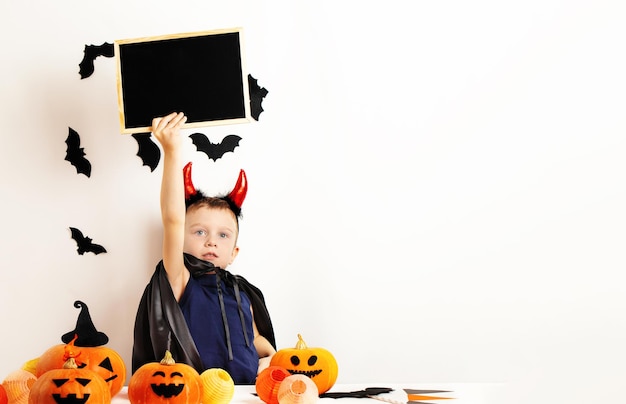 Halloween kids A funny boy in a Halloween costume and devil horns holds an empty sign in his hand Copy space