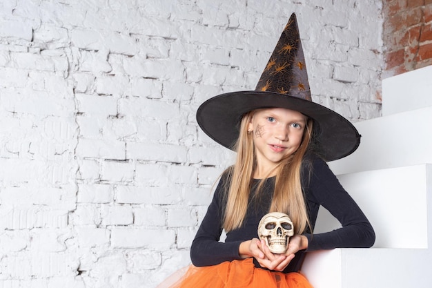 Halloween kids A smiling little blonde girl in a witch costume holding a toy skull sitting on the steps of the house