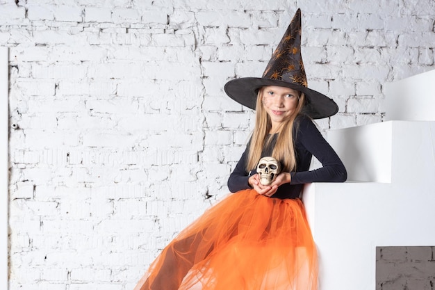 Photo halloween kids a smiling little blonde girl in a witch costume holding a toy skull sitting on the steps of the house