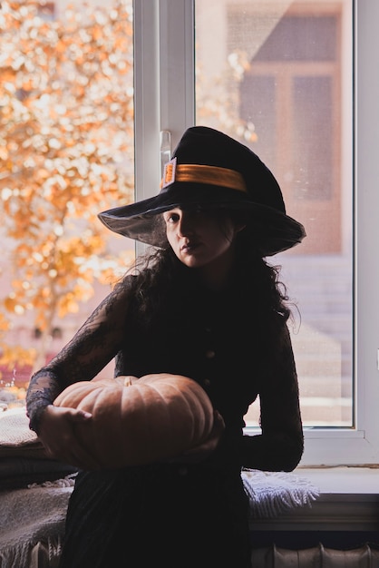 Halloween lady wearing witch cup a woman in witch hat and halloween pumpkin near the windows portrai...