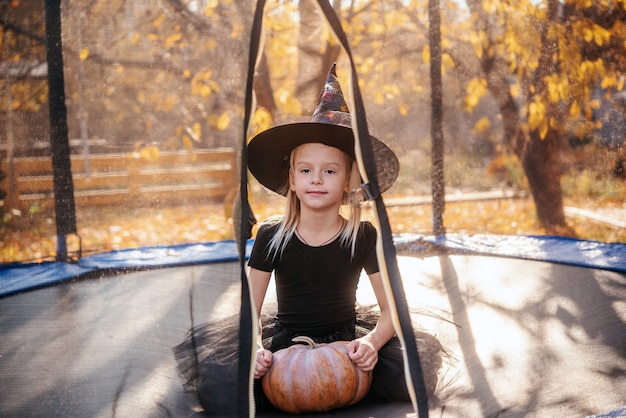 Halloween little girl in witch hat holding a pumpkin on the trampoline with yellow leaves on the background