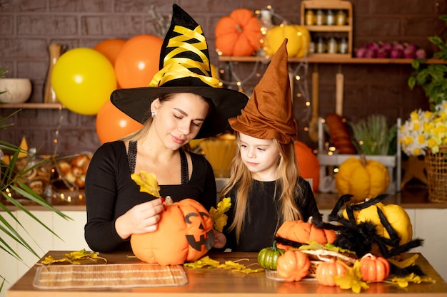 Halloween mom and daughter child in a witch costume with pumpkins and a big spider in a dark kitchen decorates a pumpkin during the Halloween celebration