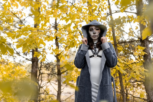 Halloween portrait A young beautiful girl in a white dress with makeup on her face in a hooded mantle creepily stands in the autumn forest The day of the Dead