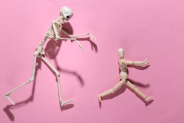 Halloween scary concept. Wooden puppet runs away from the skeleton on pink bright