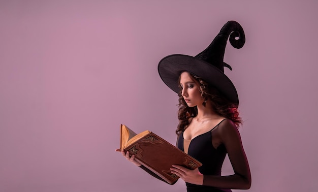 Halloween witch beautiful young woman in a witch's hat conjures practices witchcraft with an old book in her hands on a lilac background of a huge lilac moon Copy space