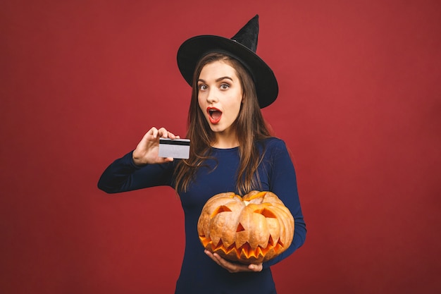 Halloween Witch with a carved Pumpkin and credit card - isolated on red background. Emotional young woman in Halloween costume. 