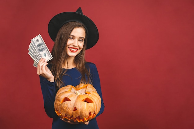 Halloween Witch with a carved Pumpkin and money - isolated on red background. Emotional young woman in Halloween costume. 