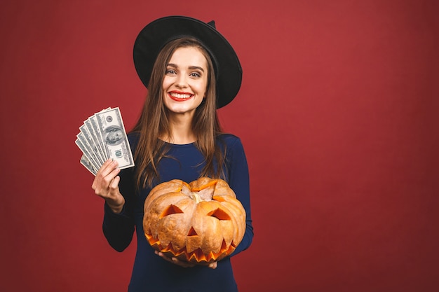 Halloween Witch with a carved Pumpkin and money - isolated on red background. Emotional young woman in Halloween costume. 