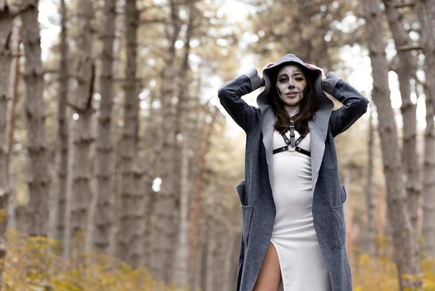 Halloween A young beautiful girl in a white dress with makeup on her face in a mantle with a hood creepily stands in the autumn forest and smiles The day of the Dead