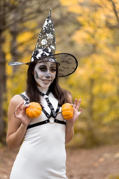 Halloween Young beautiful girl in a white dress with makeup on her face in a witch39s hat posing in the autumn forest He holds two small pumpkins in his hands and smiles The day of the Dead