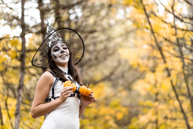 Halloween Young beautiful girl in a white dress with makeup on her face in a witch39s hat posing in the autumn forest He holds two small pumpkins in his hands and smiles The day of the Dead