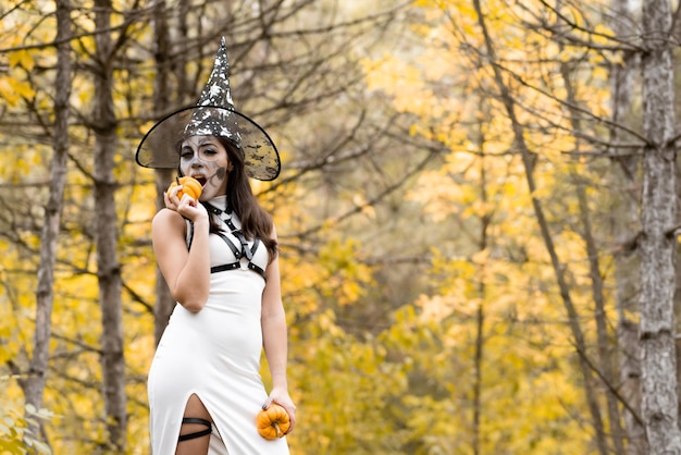 Halloween Young beautiful girl in a white dress with makeup on her face in a witch39s hat posing in the autumn forest Skeleton face makeup Bites a small pumpkin The day of the Dead